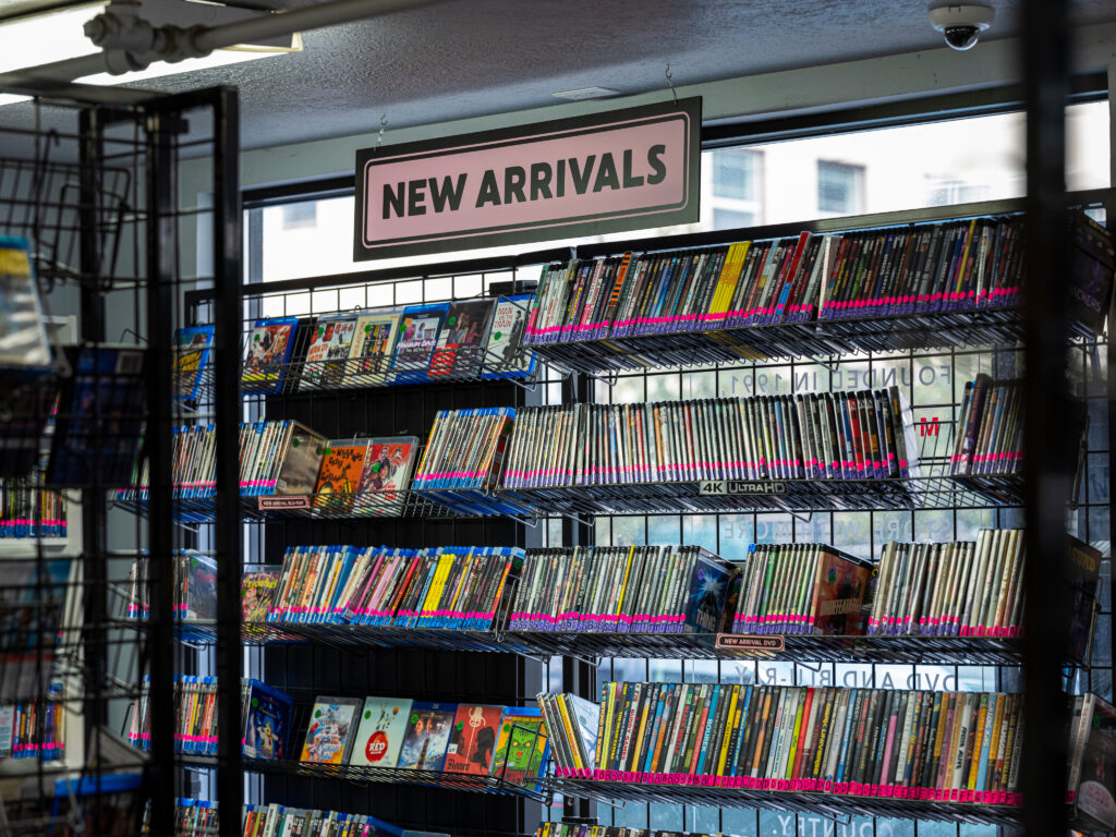Shelves filled with DVDs and Blu-rays in the 'New Arrivals' section of Movie Madness, a video rental store. A sign hangs above the display, indicating the latest titles available for rent. The scene is bright, with natural light coming in through large windows, highlighting the colorful array of movie covers.